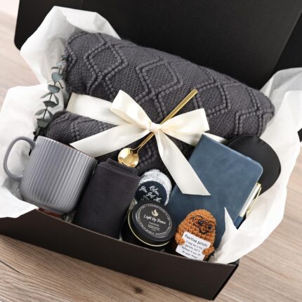 A grey and navy CASTATEO comfort care package featuring a knitted throw blanket, ceramic mug, and a small 'Positive Potato' t