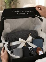 A grey and navy CASTATEO comfort care package featuring a knitted throw blanket, ceramic mug, and a small 'Positive Potato' t
