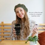 Black camera lens coffee mug with a cup sleeve and silicone straw, showcasing its unique design and accessories.