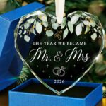 Heart-shaped glass ornament featuring "The Year We Became Mr & Mrs - 2026" in a navy blue gift box with an ivory ribbon.