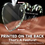 Heart-shaped glass ornament featuring "The Year We Became Mr & Mrs - 2026" in a navy blue gift box with an ivory ribbon.