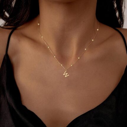 Close-up of a delicate 14K gold-plated initial 'W' necklace on a simple bead chain, worn by a woman.