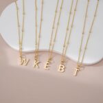 Close-up of a delicate 14K gold-plated initial 'W' necklace on a simple bead chain, worn by a woman.