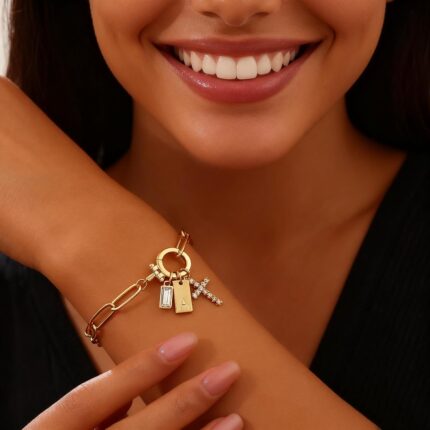 Close-up of a gold-plated paperclip link chain bracelet with a letter J initial charm and a small cross charm, on a light bac
