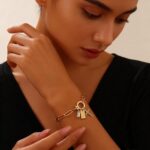 Close-up of a gold-plated paperclip link chain bracelet with a letter J initial charm and a small cross charm, on a light bac