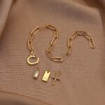 Close-up of a gold-plated paperclip link chain bracelet with a letter J initial charm and a small cross charm, on a light bac